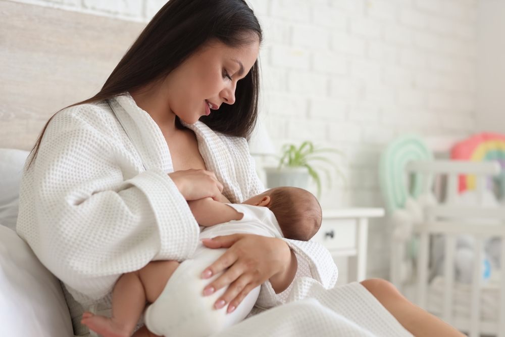 Mother in white robe breastfeeding her baby at home after breast surgery, illustrating post-operative nursing success