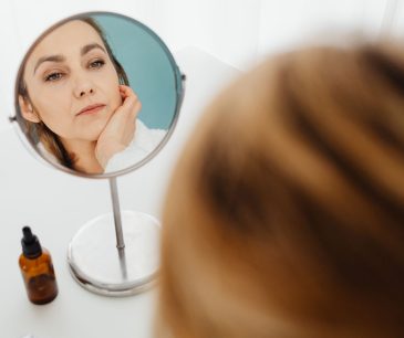 Adult woman examining her face in a mirror next to a skincare bottle