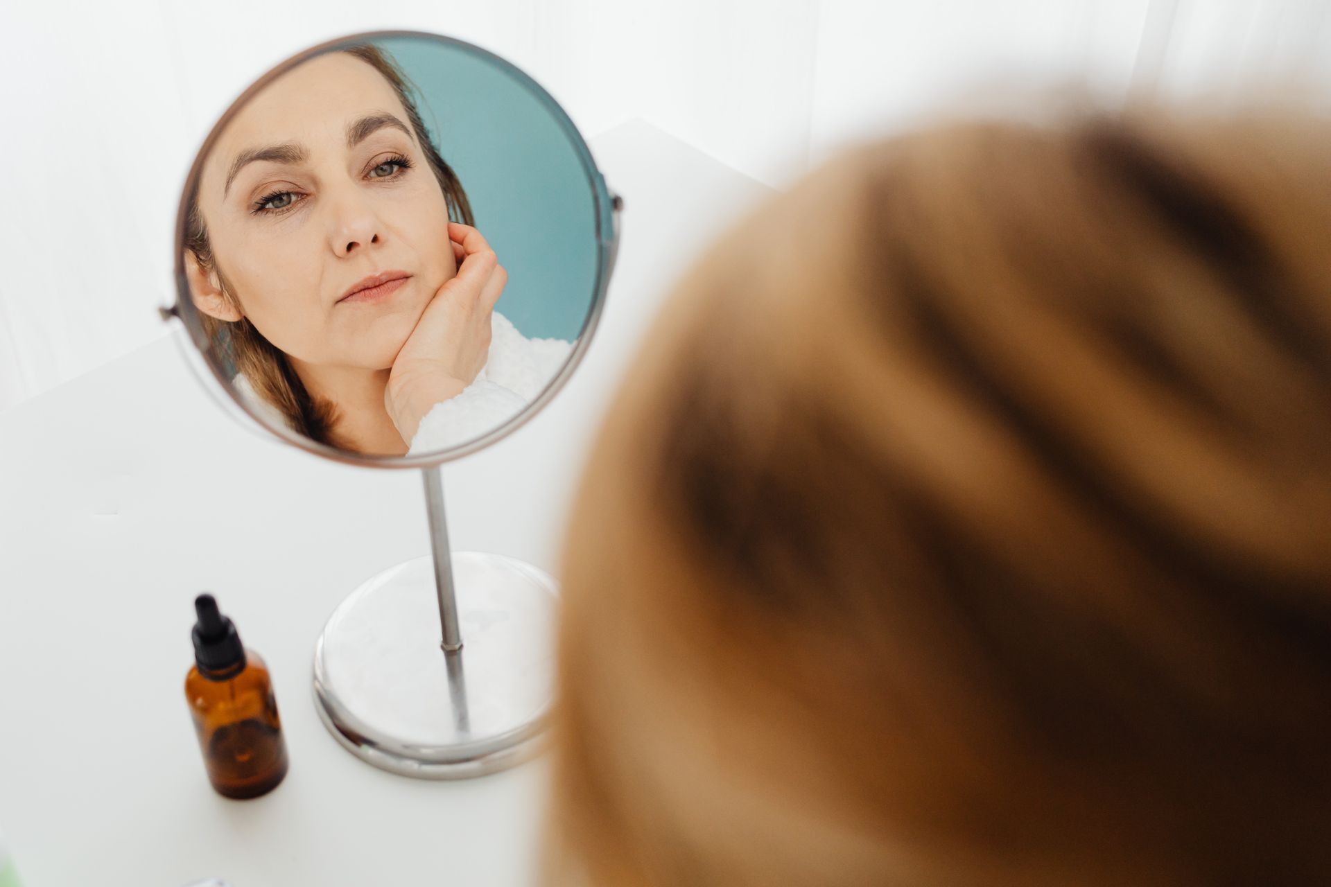 Adult woman examining her face in a mirror next to a skincare bottle
