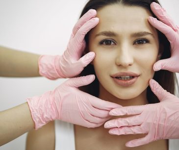 A woman receiving a facial treatment, hands in gloves gently touching her face indoors.
