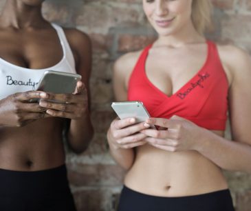 Close-up of two women in athletic wear using smartphones against a brick wall.
