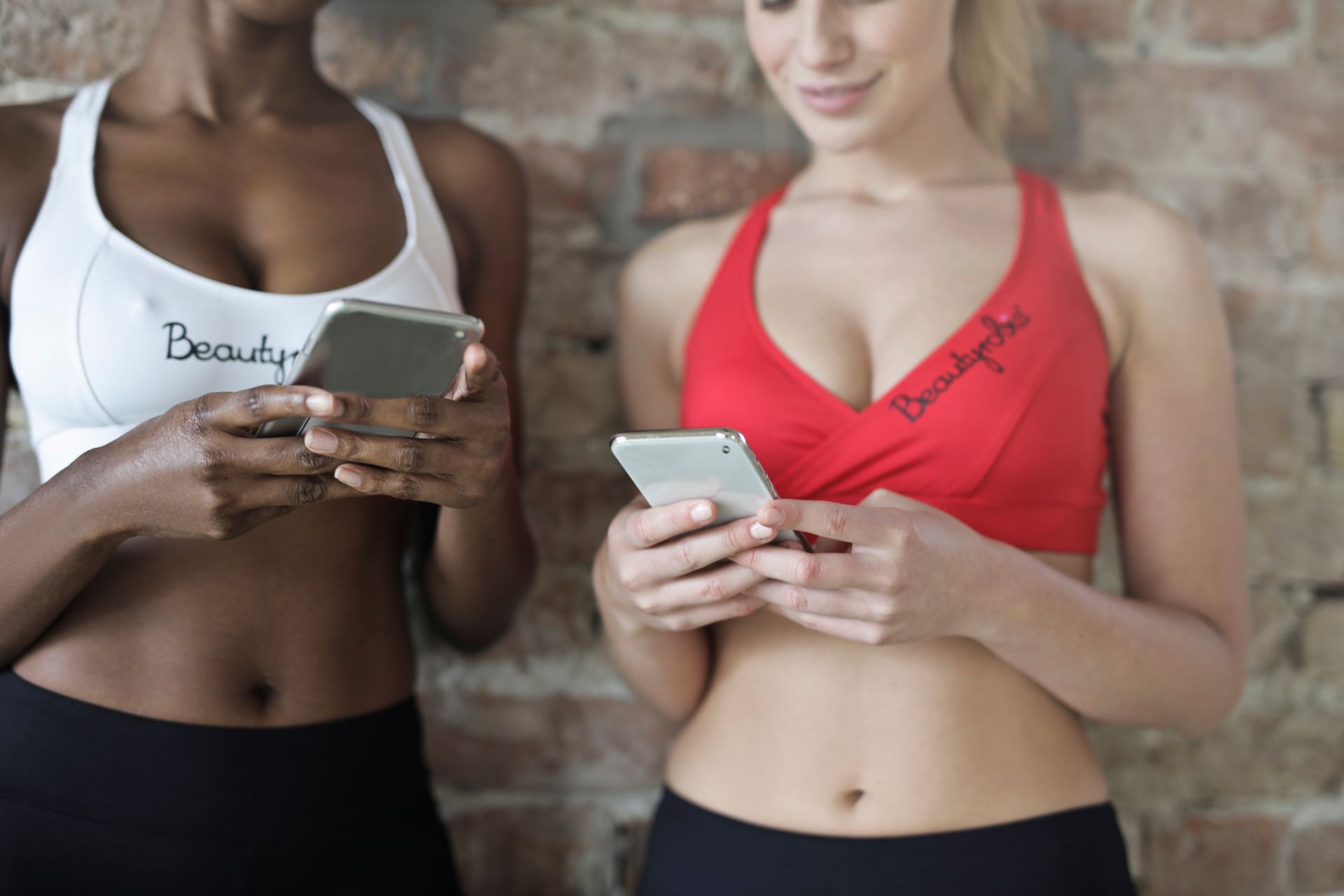 Close-up of two women in athletic wear using smartphones against a brick wall.